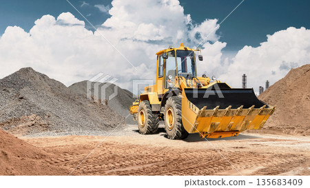 A wheel loader moves sand in its bucket on a construction site surrounded by large piles of gravel and dirt under a clear blue sky 135683409