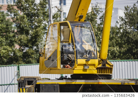 A crane operator is sitting in the cabin of a yellow crane on a busy construction site. The sun shines down on the machinery and buildings nearby 135683426