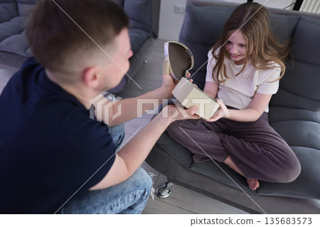 Joyful child opening a surprise gift at home with her smiling dad during a cozy afternoon Joyful child opening a surprise gift at home with her smiling dad during a cozy afternoon 135683573