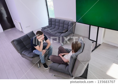 Engaging conversation takes place in a modern room with cozy gray chairs and a large screen display 135683707