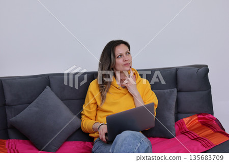 Contemplative moment of a young woman in a cozy setting while working on her laptop in a stylish living room 135683709