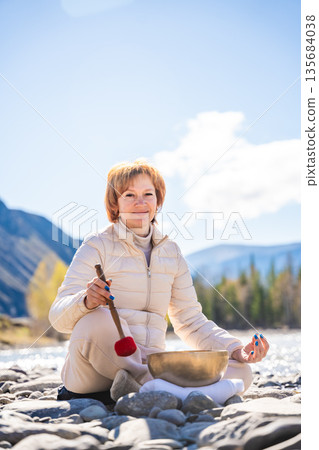 Woman performing a sound ritual with a Tibetan singing bowl by the Altai river. Concept of feminine energy, sacred vibration, and shamanic connection to the Earth Woman performing a sound ritual with a Tibetan singing bowl by the Altai river. Concept of feminine energy, sacred vibration, and shamanic connection to the Earth 135684038