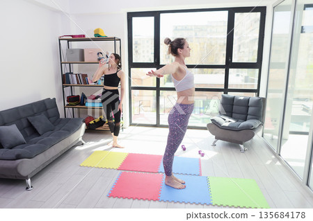 Two women practicing yoga in a bright and modern living room filled with natural light and colorful mats Two women practicing yoga in a bright and modern living room filled with natural light and colorful mats 135684178