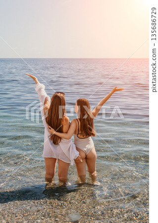 Friends beach summer two young women standing in the sea with arms raised looking at the horizon enjoying vacation freedom 135686729