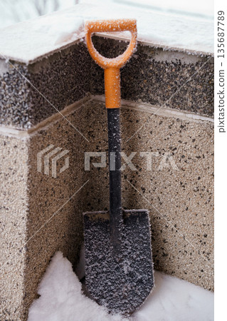 Snow-covered orange-handled shovel leaning against concrete steps in winter scene with frosty textures and cold atmosphere 135687789