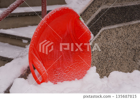 Vibrant Red Snow Sled Resting on Snow-Covered Stone Steps Amid Winter Landscape Vibrant Red Snow Sled Resting on Snow-Covered Stone Steps Amid Winter Landscape 135687815