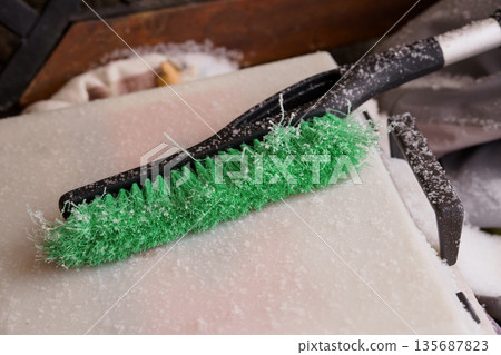 Green Ice Scraper Brush Covered in Snow on Car Windshield During Winter Weather 135687823