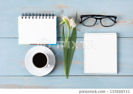 A blue wooden desk with blank notebooks, glasses, a pen, gladiolus, and coffee. Morning coffee cup. A blue wooden desk with blank notebooks, glasses, a pen, gladiolus, and coffee. Morning coffee cup. 135688531