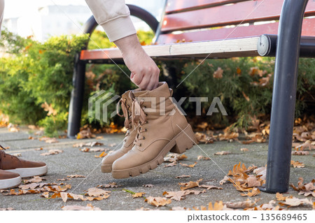 Young person's hand carefully holds fashionable tan leather combat boots featuring a thick lug sole, representing versatile autumnal footwear choice for comfortable casual outdoor walks 135689465