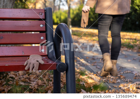 A single beige glove sits abandoned on a weathered wooden park bench, surrounded by vibrant fallen autumn leaves, representing themes of loss, departure, and the passage of time A single beige glove sits abandoned on a weathered wooden park bench, surrounded by vibrant fallen autumn leaves, representing themes of loss, departure, and the passage of time 135689474