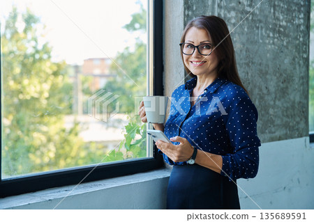 Middle aged beautiful businesswoman with smartphone, cup, near window 135689591