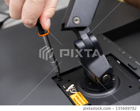 Hand carefully attaching a computer display to an adjustable desk mount using a screwdriver, symbolizing precise assembly and technical installation Hand carefully attaching a computer display to an adjustable desk mount using a screwdriver, symbolizing precise assembly and technical installation 135689782