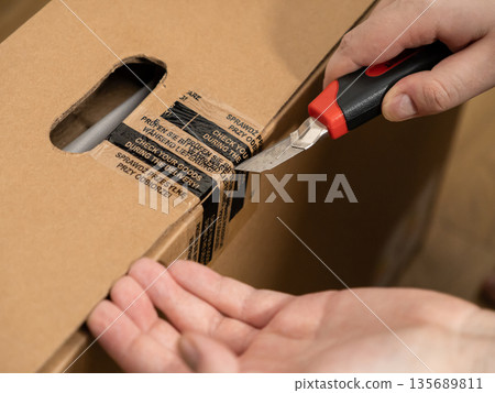 Person's hand uses a utility knife to open a cardboard box, with a visible label encouraging careful goods inspection during delivery Person's hand uses a utility knife to open a cardboard box, with a visible label encouraging careful goods inspection during delivery 135689811