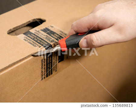 Close-up of a person carefully cutting the sealed brown cardboard shipping box with a sharp utility knife, emphasizing secure delivery and package inspection for consumer safety 135689813