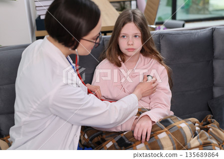 Doctor checks health of a young girl wrapped in a blanket during a home visit in a cozy living room setting on a chilly day Doctor checks health of a young girl wrapped in a blanket during a home visit in a cozy living room setting on a chilly day 135689864