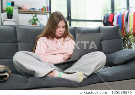 Girl focused on her laptop while sitting comfortably in a cozy living room with colorful clothes in the background 135689870