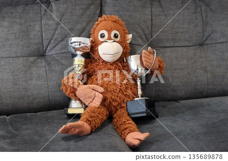 A stuffed monkey is sitting on a couch holding two cups and two trophies A stuffed monkey is sitting on a couch holding two cups and two trophies 135689878