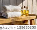 Sunlit towels on rustic bench, folded towels near glass oil bottles, warm window shadows draw texture on wood, towels promise clean comfort and spa freshness in cozy morning light glow 135690305
