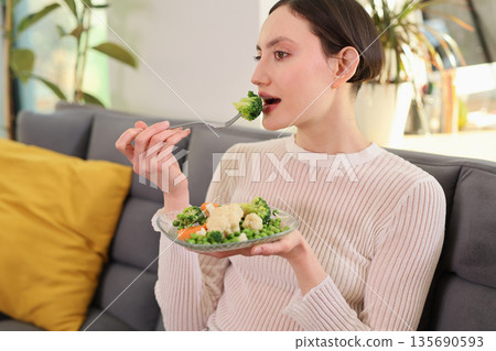 Eating a healthy plate of fresh vegetables while relaxing on a cozy sofa during a sunny afternoon indoors Eating a healthy plate of fresh vegetables while relaxing on a cozy sofa during a sunny afternoon indoors 135690593