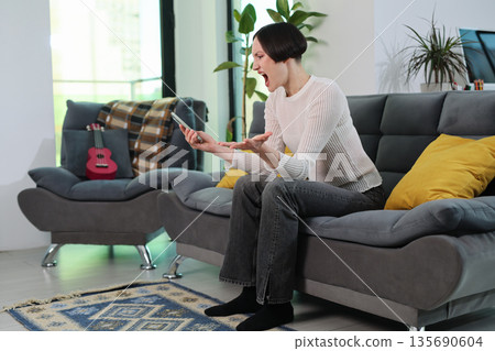 Frustration visible as a young woman expresses her emotions while using a smartphone in a cozy living room Frustration visible as a young woman expresses her emotions while using a smartphone in a cozy living room 135690604