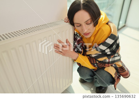 Woman in cozy sweater hugging a radiator while staying warm in a sunlit room during cold weather 135690632