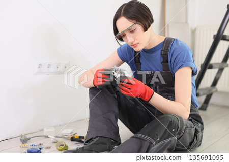 Focused electrician examines a complex circuit component while sitting on a wooden floor in a well-lit workshop Focused electrician examines a complex circuit component while sitting on a wooden floor in a well-lit workshop 135691095