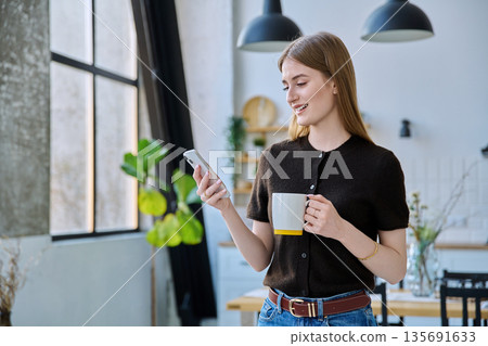 Young woman with cup of drink using smartphone standing at home Young woman with cup of drink using smartphone standing at home 135691633
