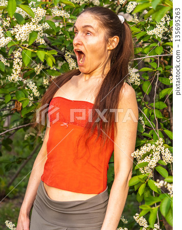 Young woman sneezes in the park against the background of a flowering tree. Allergy to pollen concept. Young woman sneezes in the park against the background of a flowering tree. Allergy to pollen concept. 135695263