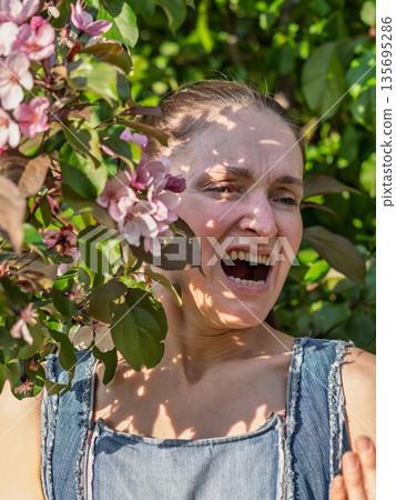 Young woman sneezes in the park against the background of a flowering tree. Allergy to pollen concept. Young woman sneezes in the park against the background of a flowering tree. Allergy to pollen concept. 135695286
