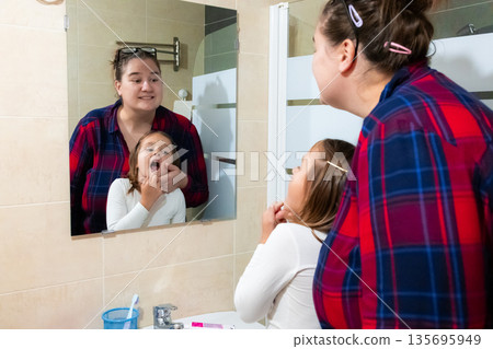 Mother and daughter brushing teeth together in bathroom mirror reflection Mother and daughter brushing teeth together in bathroom mirror reflection 135695949