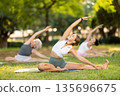 Portrait of caucasian woman sitting on sports mat and making yoga meditation in lotus pose and hands up with group together in park at daytime 135696675