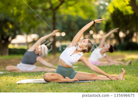 Portrait of caucasian woman sitting on sports mat and making yoga meditation in lotus pose and hands up with group together in park at daytime 135696675