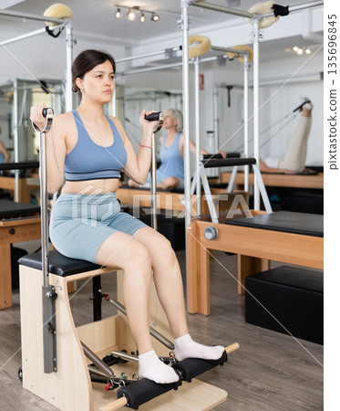 Slim woman engaging in pilates training on fitness chair in exercise room during workout session Slim woman engaging in pilates training on fitness chair in exercise room during workout session 135696845