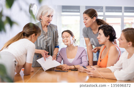 Women discuss documents sitting at table in office Women discuss documents sitting at table in office 135696872