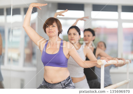 Women doing ballet at group amateurs training session in the studio performs demi plie near the ballet barre, standing in ballet stance 135696970