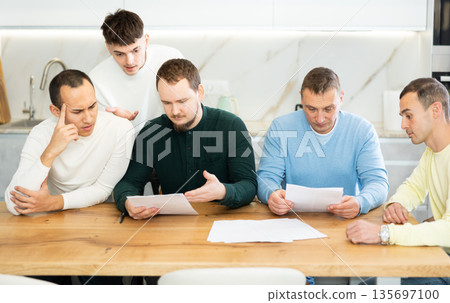 Friends reading financial documents and trying to solve problems together while sitting at table in their kitchen Friends reading financial documents and trying to solve problems together while sitting at table in their kitchen 135697100