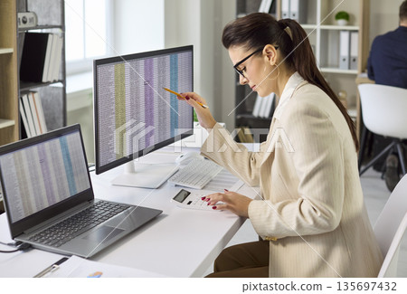 Female accountant analyzing spreadsheets on dual monitors in modern office audit Female accountant analyzing spreadsheets on dual monitors in modern office audit 135697432