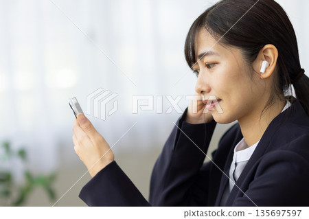 A young woman in a suit wearing wireless earphones and looking at her smartphone at a table by the window A young woman in a suit wearing wireless earphones and looking at her smartphone at a table by the window 135697597