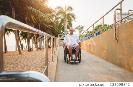 Disabled man in a wheelchair moves on a ramp to the beach. Disabled man in a wheelchair moves on a ramp to the beach. 135699842