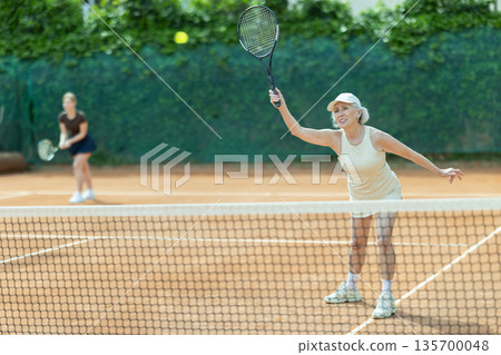 Elderly woman playing doubles tennis on court 135700048