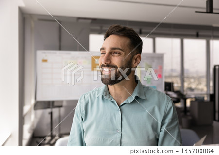 Businessman standing in office, looks out window with contented smile Businessman standing in office, looks out window with contented smile 135700084