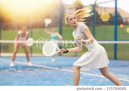 Woman actively playing paddle tennis on a court in a park against the background of glare Woman actively playing paddle tennis on a court in a park against the background of glare 135700089