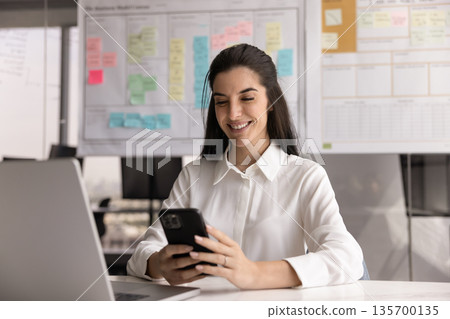 Professional woman sits at desk using smartphone with contented smile Professional woman sits at desk using smartphone with contented smile 135700135