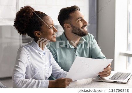 Teammates looking into distance with contented smile in workplace Teammates looking into distance with contented smile in workplace 135700228