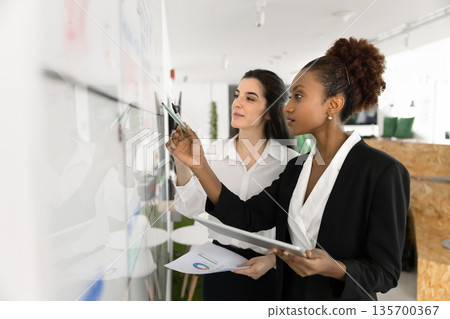 Two women colleagues analyzing data standing next to white board Two women colleagues analyzing data standing next to white board 135700367