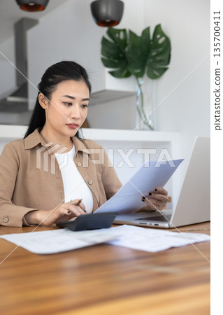 Asian woman managing personal finances seated at table at home Asian woman managing personal finances seated at table at home 135700411