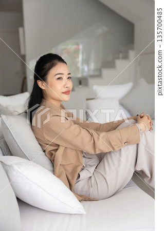 Thoughtful Asian woman dressed casually sits on cozy white couch Thoughtful Asian woman dressed casually sits on cozy white couch 135700485