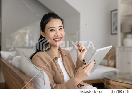 Woman looking at camera seated on cozy sofa with tablet 135700506