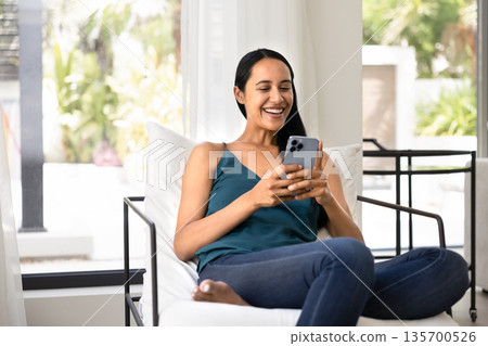Happy indigenous woman typing on mobile phone in home armchair 135700526