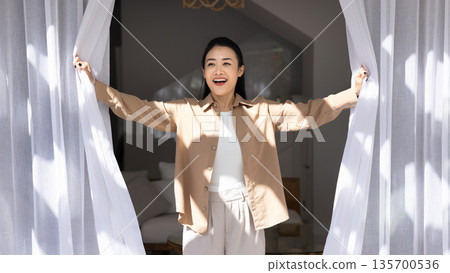 Excited Asian woman opening white curtains lets sunlight into room 135700536